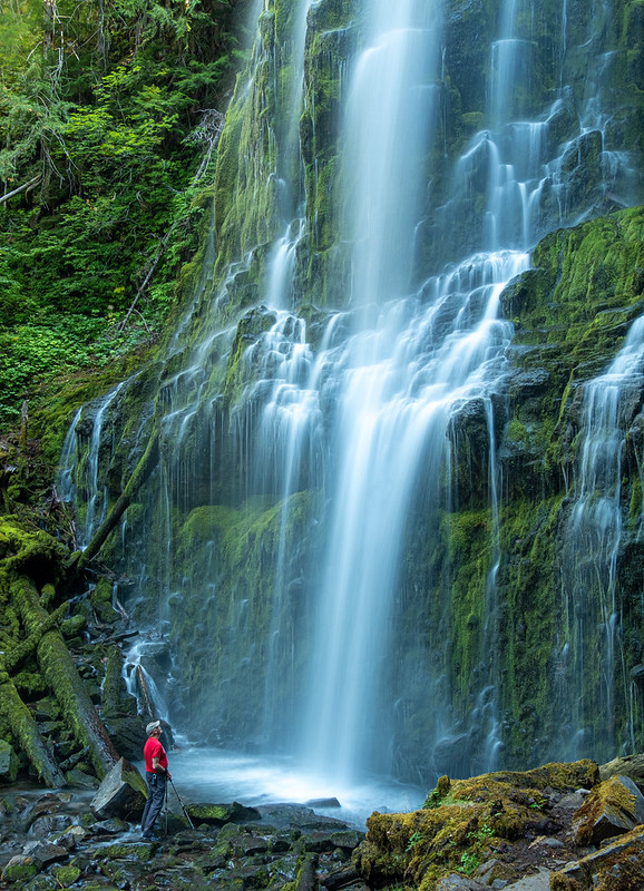 Water cascades over mossy cliff and rocks. A man in a red shirt is at the bottom of the falls staring up. He looks tiny.