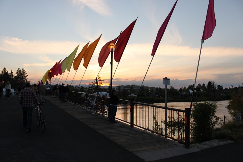 A footbridge goes over the river in Bend at sunset. People are walking across it.