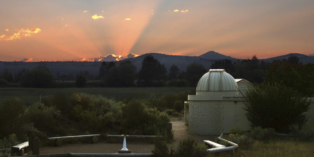 The observatory in Sunriver at sunset. Rays of sun peak out over low mountains in the distance. There are trees, a field, and desert brush around the small white observatory building. There are also wooden benches in a circle outside the building.