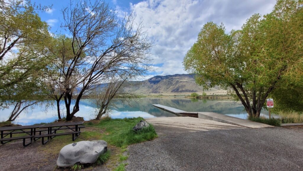 A dock goes down into Ana Reservoir on a nice day. There are a few trees, some grass, and a picnic bench on the shore of the still waters of the reservoir.