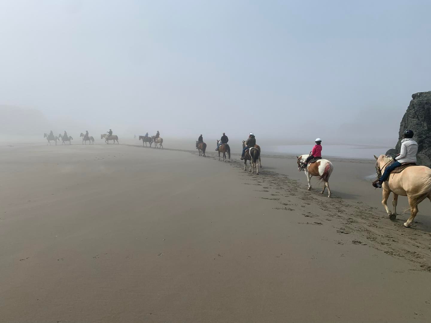 Photo via the Bandon Beach Riding Stables Facebook page. A group of people rides horses down the beach in the fog in Bandon, Oregon.
