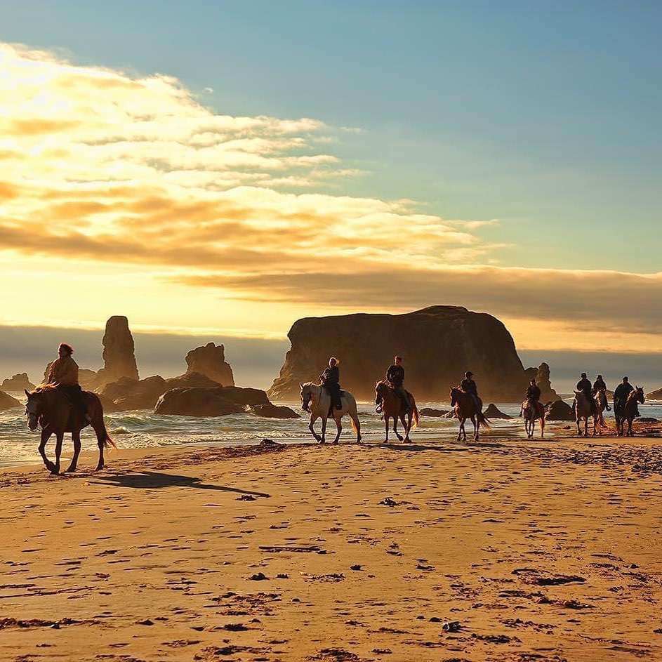 Photo via Bandon Beach Riding Stables website. A group of people riding horses down the coast in Bandon Oregon in front of seastacks.