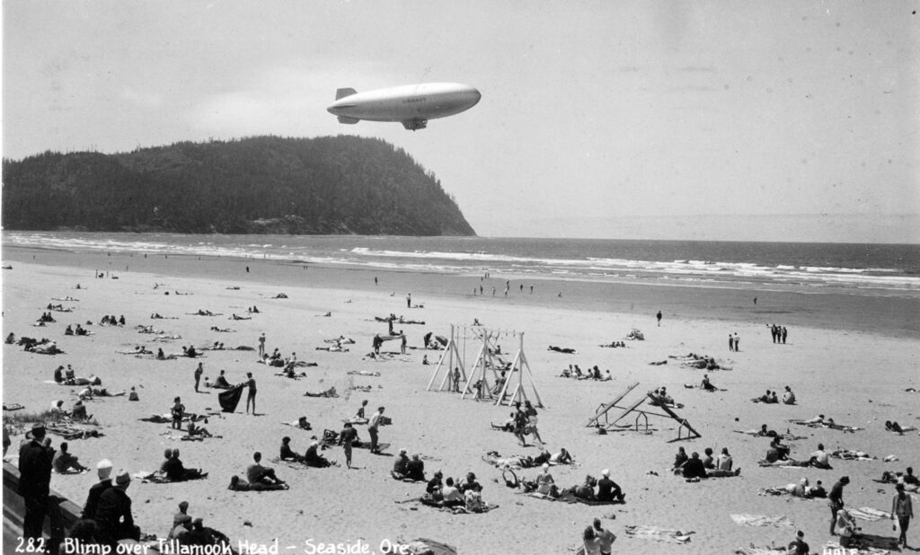 A black and white photo of a K ship flying over the water in Seaside Oregon. Beachgoers look on. tillamook air museum, planes, WWII, history, family things to do, oregon coast