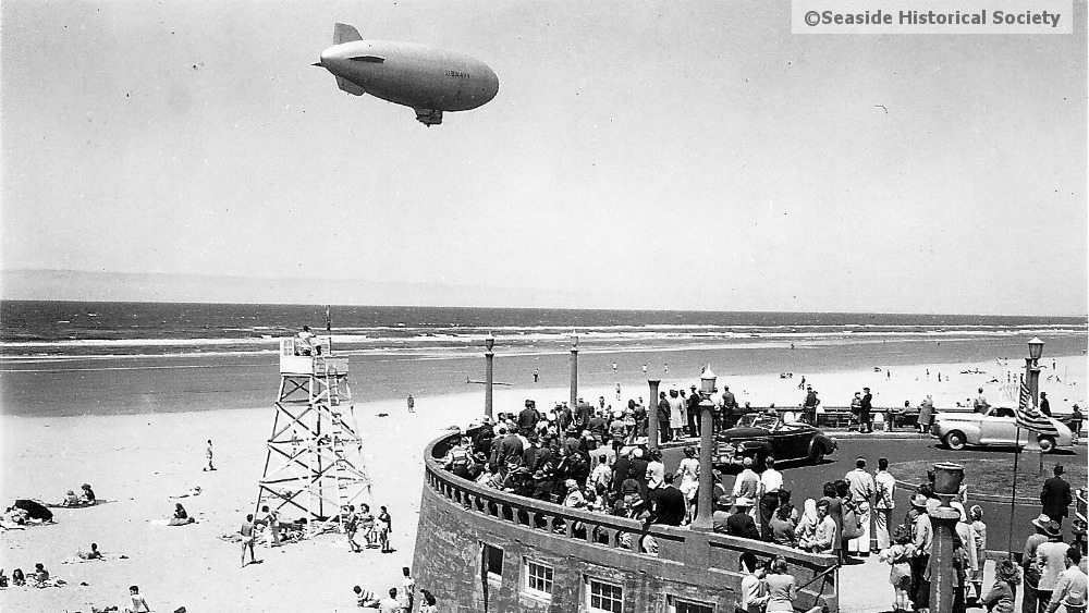 A black and white photo of a K ship flying over the water in Seaside Oregon. Beachgoers look on. tillamook air museum, planes, WWII, history, family things to do, oregon coast