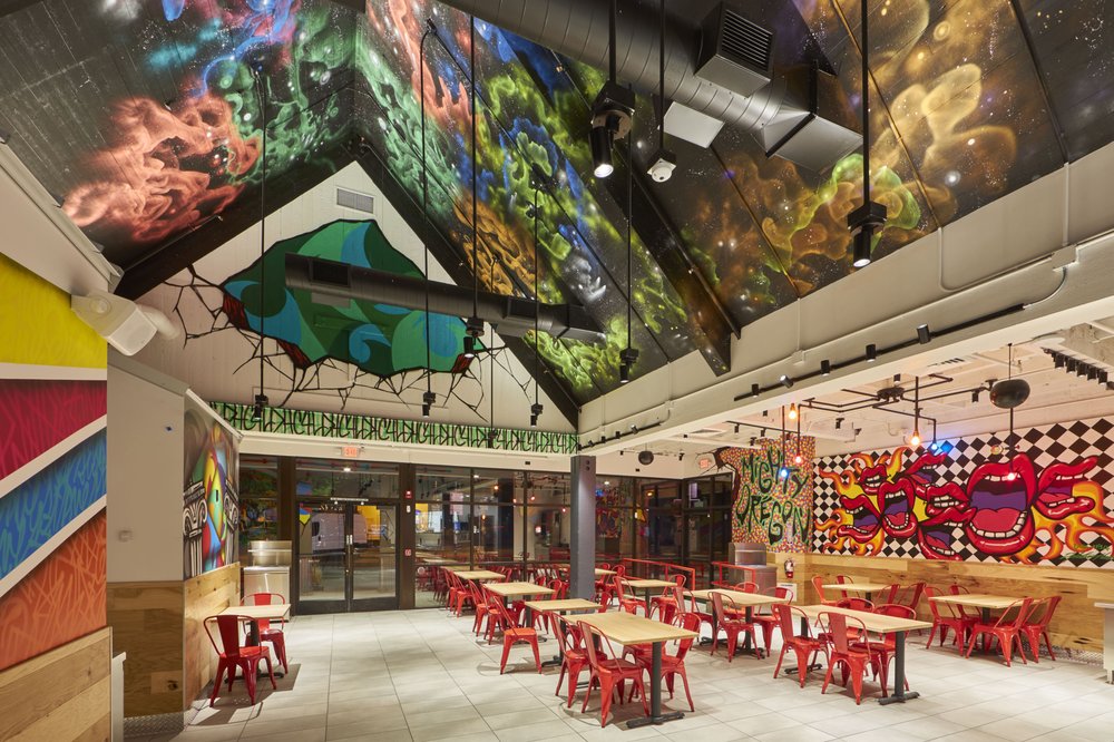 The interior of Dave's Hot Chicken in Eugene, Oregon. There's a vaulted ceiling with graffiti art, red chairs and wooden tables, and graffiti art on the walls.
