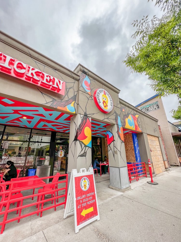 The exterior of Dave's Hot Chicken in Eugene, Oregon at daytime. There is red and blue graffiti style paint on the ceiling, bright red chairs and tables outside, and the whole place looks like it has a hip fun vibe.