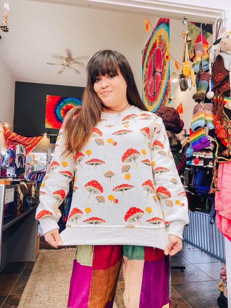 A woman shows off a white sweatshirt that she's wearing with red mushrooms, and other color mushrooms on it. A variety of knit socks hang in the background.
