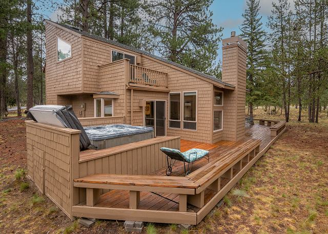 The exterior of Duckpond #2. It's a brown two story house with a large wrap around wooden porch and a hot tub. There are pine trees in the background.