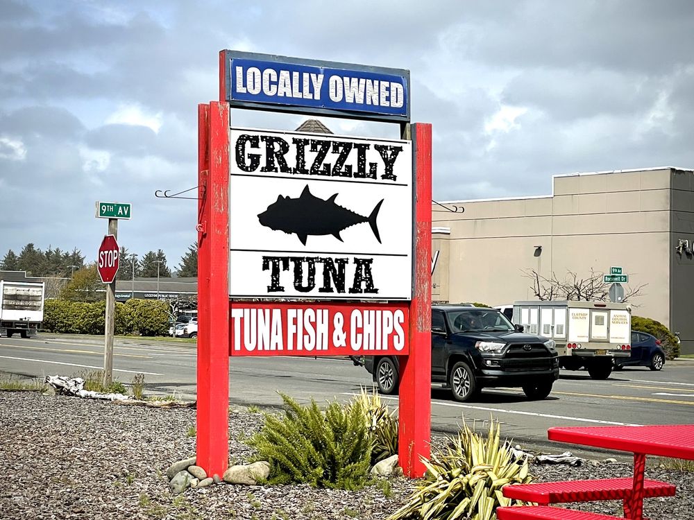 Photo by Kenny C. via Yelp. The sign for Grizzly Tuna. The sign is white with black words and a black silhouette of a tuna fish. It says "Locally Owned, Tuna Fish & Chips." The sign is held up by two bright red supports.