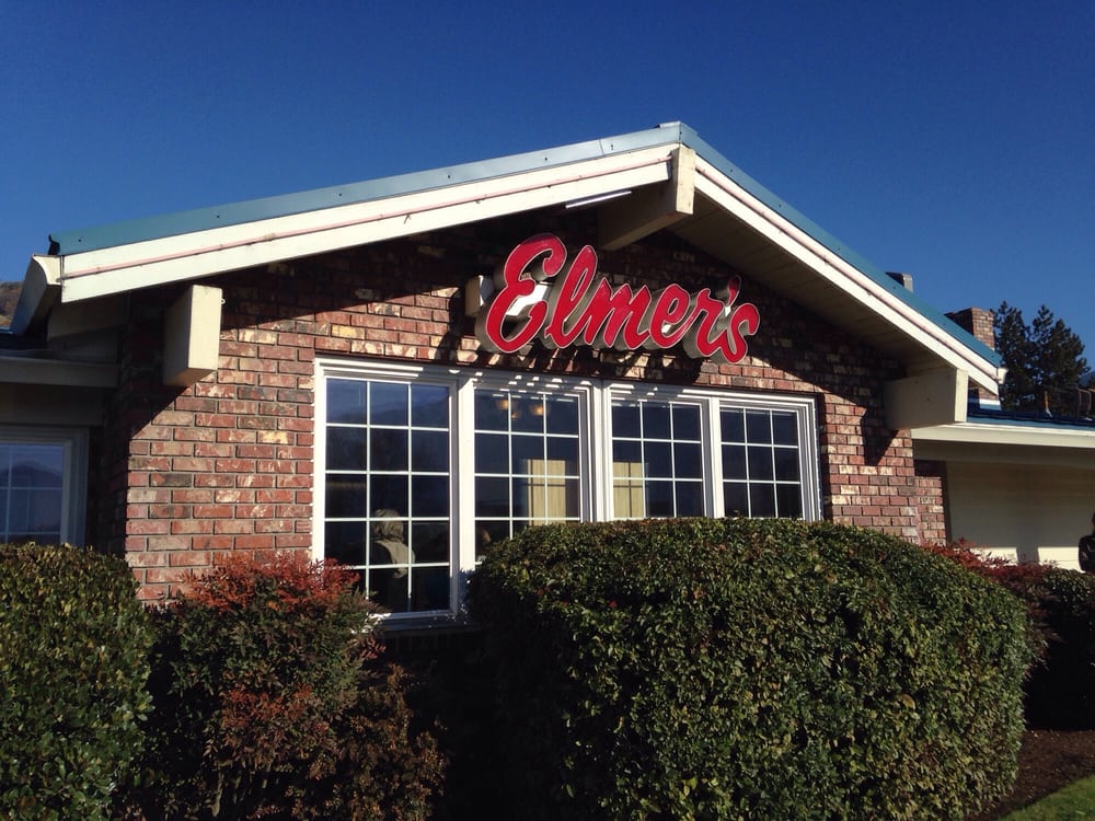 The outside of an Elmer's Restaurant. The restaurant is brick with bushes in front of a large window. There's a red Elmer's sign above the windows on the side of the building. The sign is in cursive.