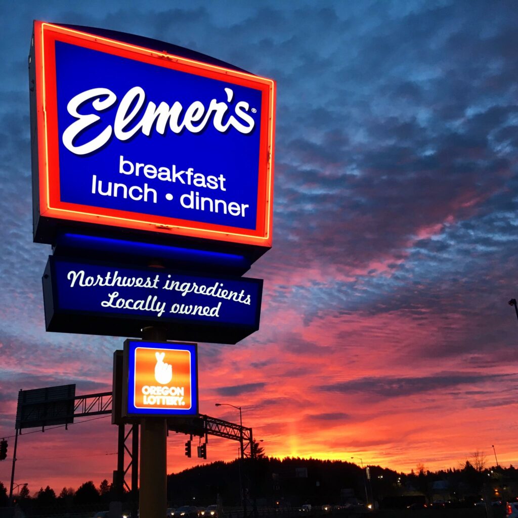 A blue and red Elmer's sign lit up as the sky grows dark with bright red, orange, pink and yellow colors from the sunset in the background. There are interesting looking clouds in the sky behind the sign. The sign reads: Elmer's - Breakfast, lunch, dinner.