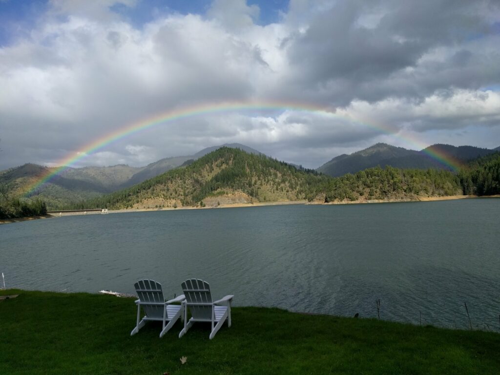 Two white chairs in front of Applegate Lake. A rainbow is across the lake on a cloudy day in front of some mountains.