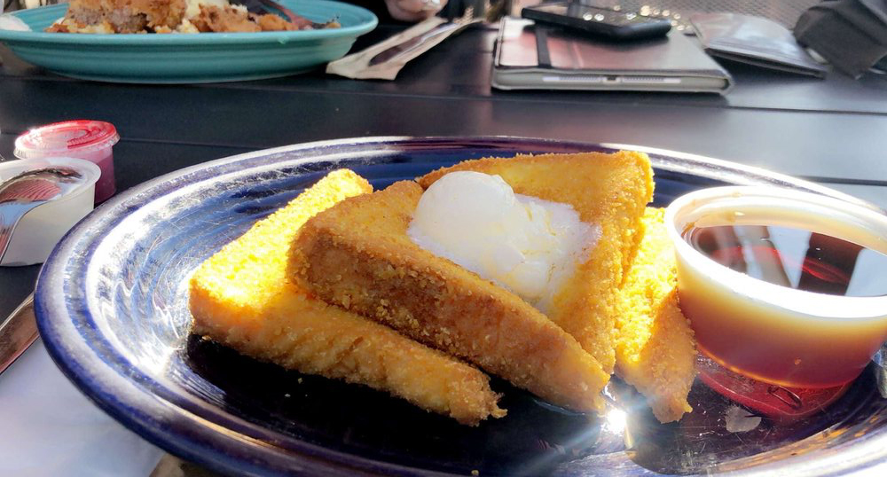 The corn field French toast on a dark blue plate with an even brighter blue plate filled with food in the background.