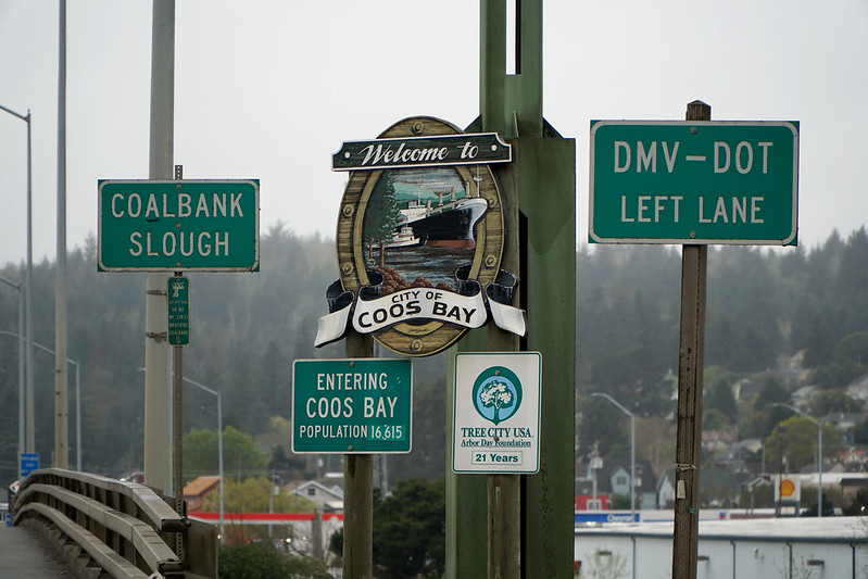 The Welcome To Coos Bay Sign on a rainy day. It's surrounded by other road signs.
