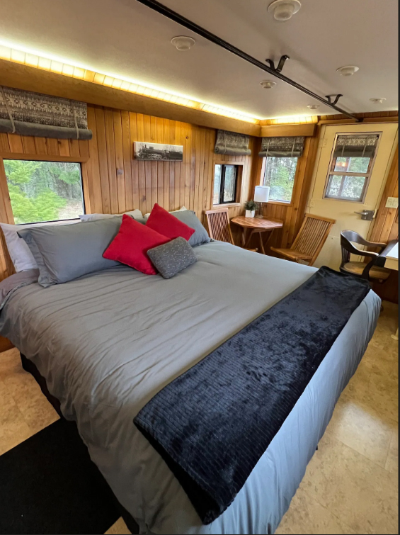 A large bed with gray and black blankets, and gray, red and white pillows. The walls are wood paneled.