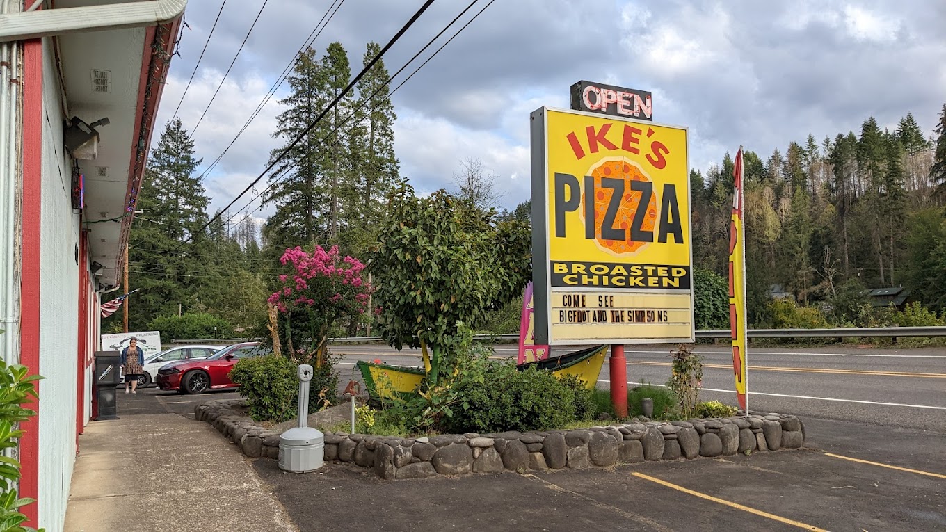 Photo by Mr. Tutthole via Google Local. A bright yellow Ike's Pizza sign surrounded by plants.