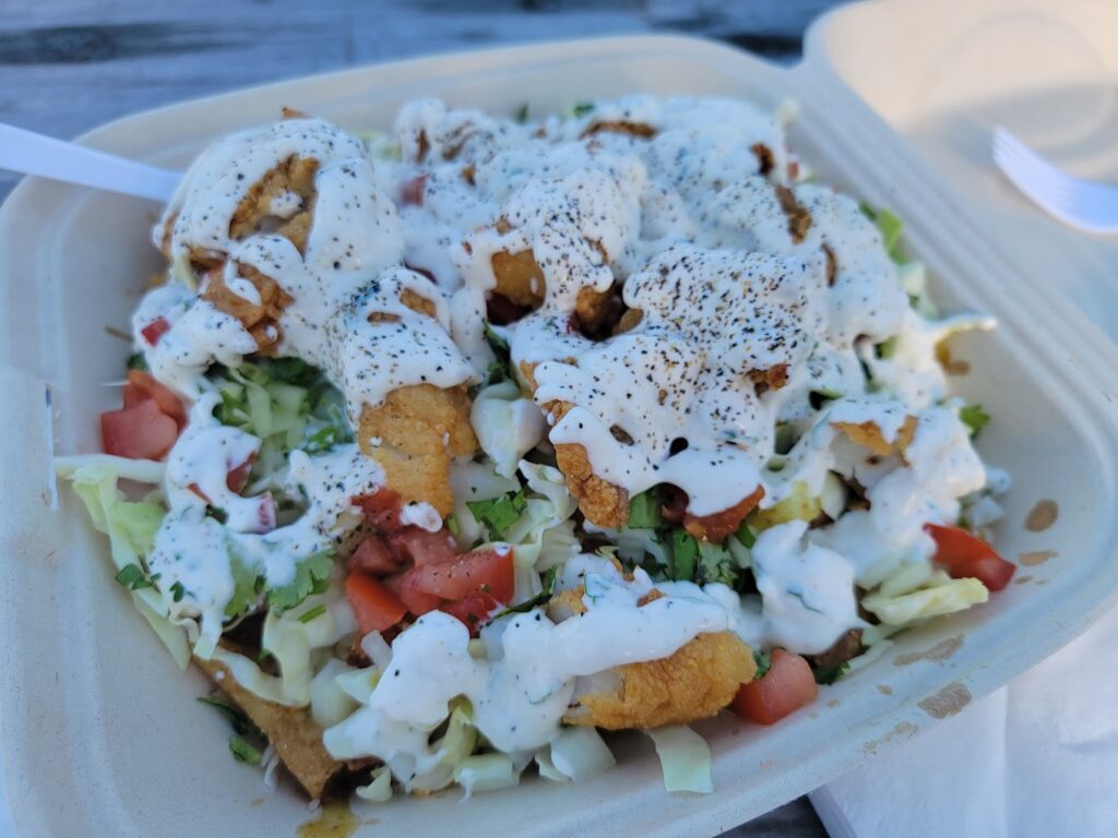A Styrofoam container with fried seafood, white sauce, seasoning, coleslaw and a lot of fresh toppings.
