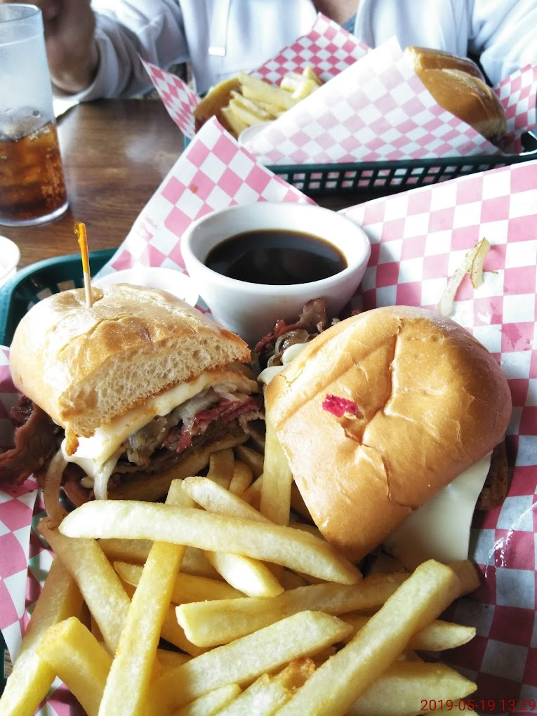 A super delicious looking sandwich with steak and cheese and what looks to be au jus dip. There are fries with it on a red and white checkered paper wrapper.