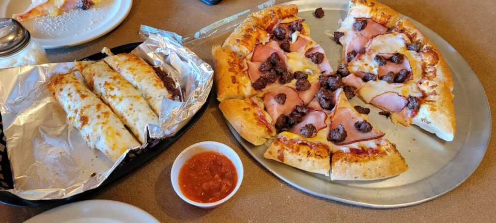 A asty pizza and breadsticks and a cup of marinara sauce for dipping.