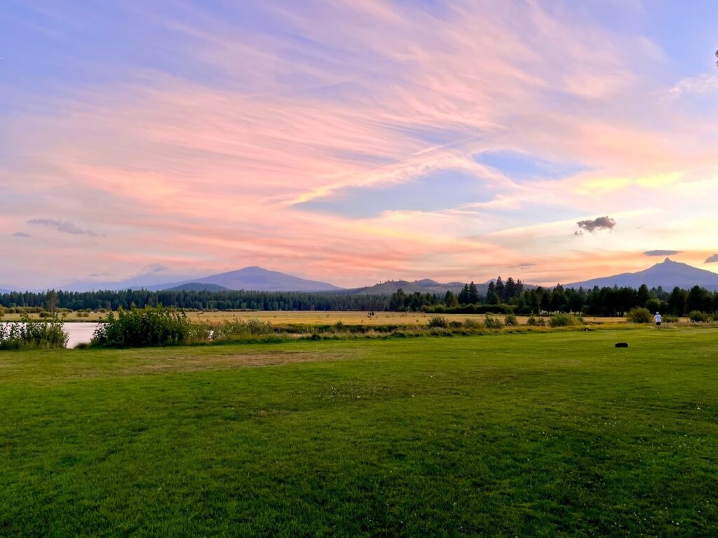 A gorgeous view of meadows and a mountain in the distance at sunset. The sunset is showing off soft pink and blue hues with fluffy and wispy clouds.