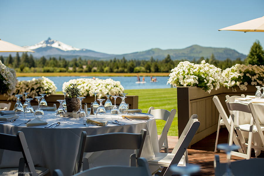 A round dining table covered in a white cloth. There are white chairs, flowers on the table, and a lake in the background along with a snow covered mountain and a ponderosa pine forest.
