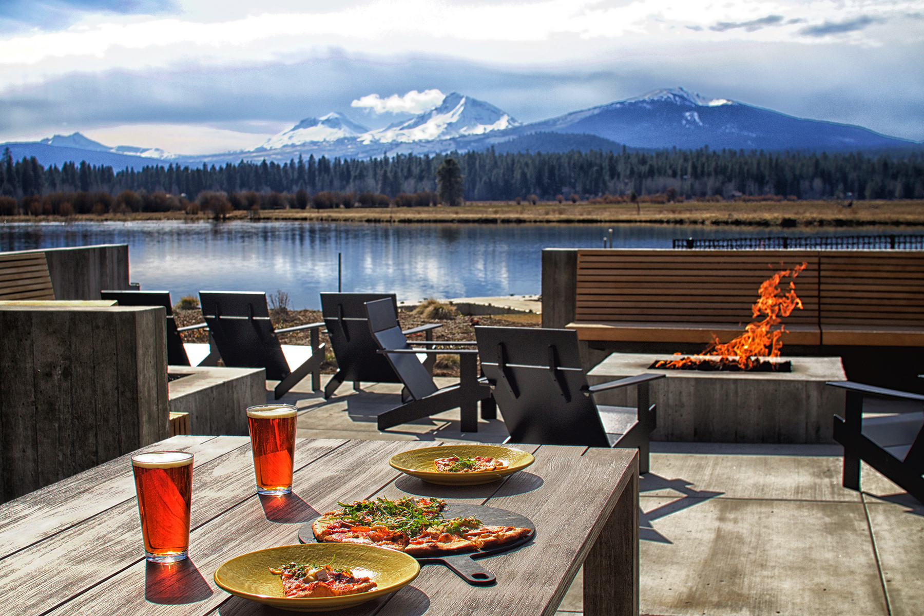 Photo via Black Butte Ranch Facebook page. Food sits on outdoor tables by a modern fire pit. In the background you can see a lake, and on the other side of the lake, there's a forest and gorgeous mountains covered in snow. It's a cloudy day.