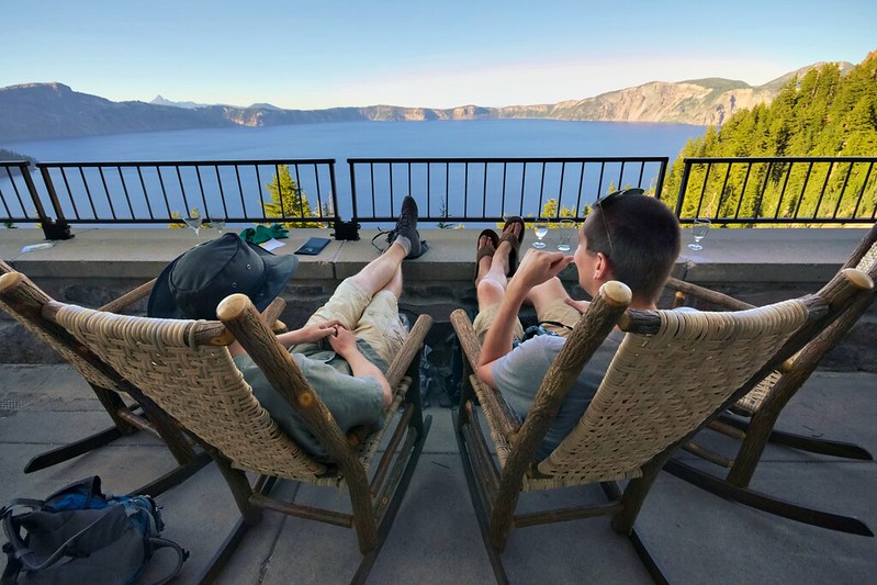 Two people sit in chairs on the deck overlooking Crater Lake. They look relaxed.