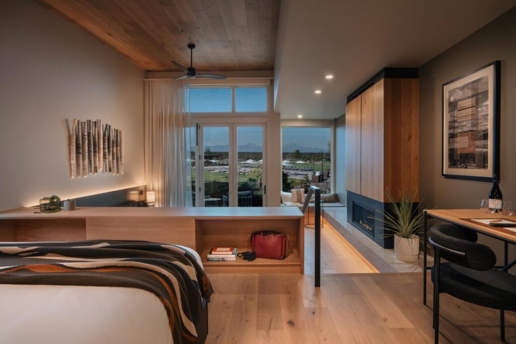 The interior of one of the bungalows. There are soft wood tones on the floors, walls, ceiling, and furniture. There's a dark gray couch and round black tables and a fireplace. The bed has white bedding and a western themed blanket. It's very pretty.