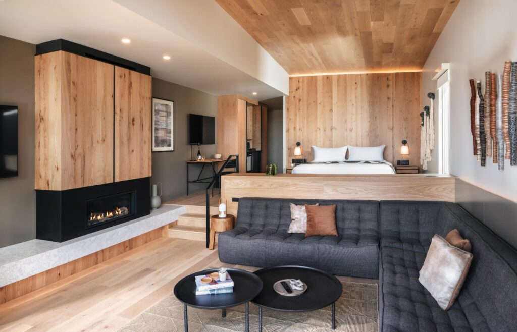 The interior of one of the bungalows. There are soft wood tones on the floors, walls, ceiling, and furniture. There's a dark gray couch and round black tables and a fireplace. The bed has white bedding.
