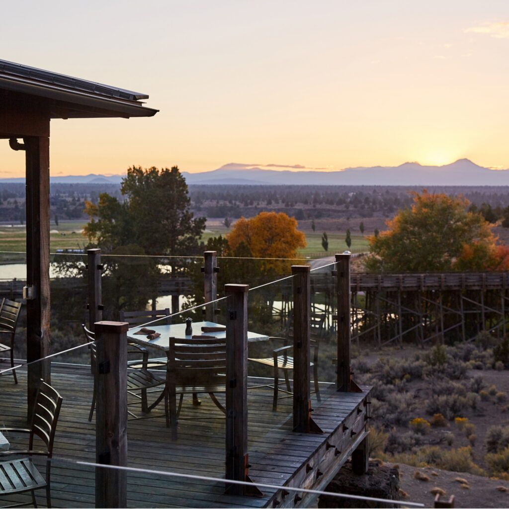 A patio with tables and chairs at Brasada Ranch overlooking the high desert. There are mountains in the distance, and the sun is setting behind the mountains, casting them in a gray blue silhouette with a slight pink hue.