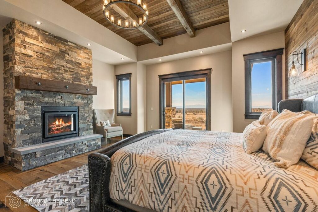 One of the rooms you can rent at Brasada Ranch. There's a stone fireplace, a western style chandelier, and a luxurious bed with very pretty western style bedding. There are windows overlooking the high desert.
