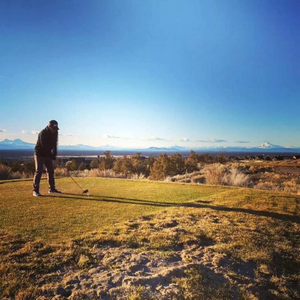 A man tees off on the Brasada Golf Course. It's a sunny day with clear blue skies, and there are mountains in the distance.