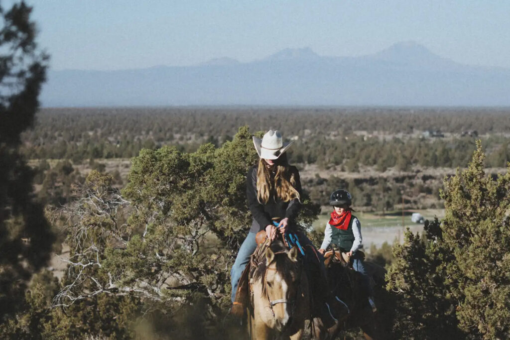 Two people ride horses in Oregon's high desert at Brasada Ranch. They're surrounded by sage and there are mountains in the background. One of the girls riding horses is wearing a white cowboy hat.