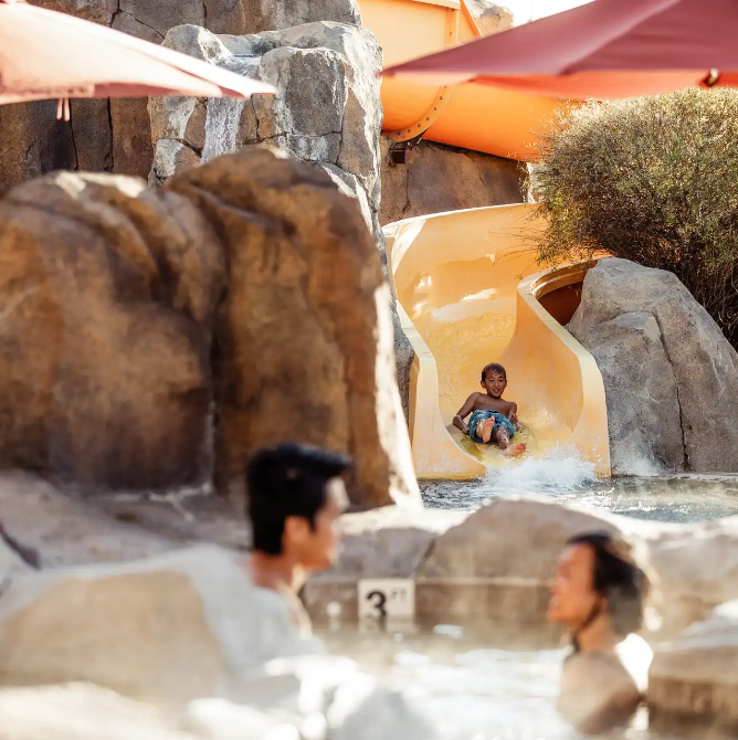 A child slides down a big yellow water slide into an outdoor pool. Two adults are in the pool smiling at each other.