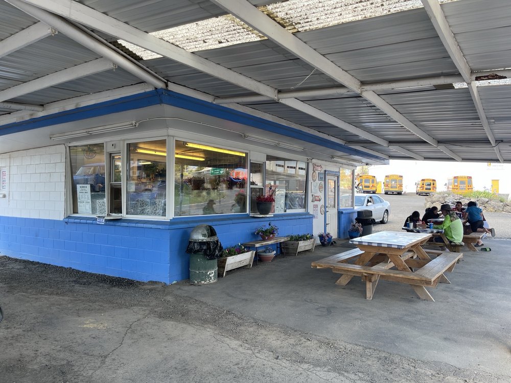 Photo by Brad T. via Yelp. The Drive In as seen from under the overhang. The building is blue and white, and there are picnic tables to eat at underneath the overhang where it's shaded.