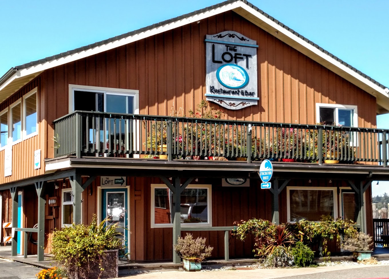 Photo by David Kennedy via Google Local. The outside of The Loft Restaurant And Bar. It's a brown building with a patio and potted plants outside. The sign is gray and blue and has a beachy vibe.