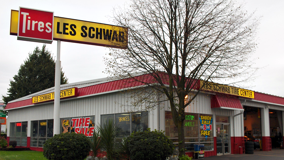 Photo via Les Schwab Tire Center via Google Local. The exterior of a Les Schwab tire store. The building is white with a red roof, and a yellow sign.