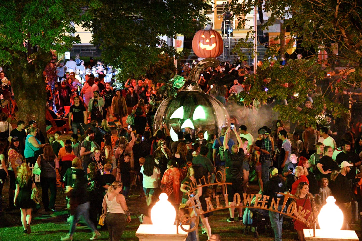 Photo via the Spirit Of Halloweentown Facebook A crowd gathers in front of a massive pumpkin with green glowing eyes at night.