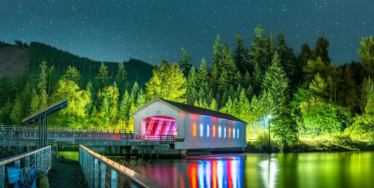 Photo by TRΞLLP via Google Local. A nighttime shot of the Lowell Covered Bridge. The bridge is white and has many colorful lights coming out from inside it. Stars hang in the sky, nad green water sits below. Trees stand tall above the bridge in the background as a path leads to it in the foreground.