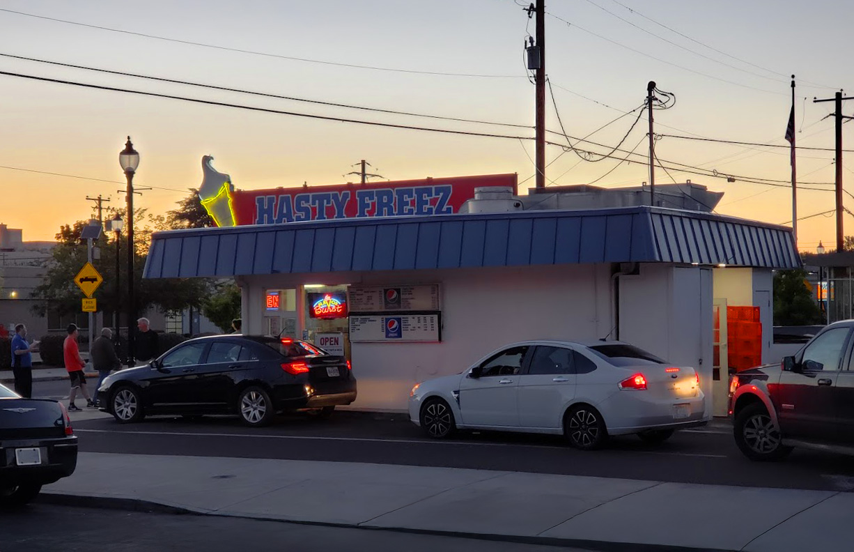 Photo by William Broadhead via Google Local. The outside of Hasty Freez at night time. There's a line of cars at the drive up window. It's a small white building with a blue roof, and a red sign with blue letters on top declaring 'Hasty Freez'. On the sign is a white and yellow neon ice cream cone.