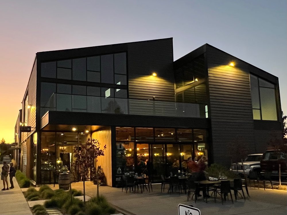 Photo by James H. via Yelp. The outside of Rancher Butcher Chef in Bend, Oregon. It's a modern, sleek black building with a lot of windows and warm lighting outside. There are tables outside to eat at.