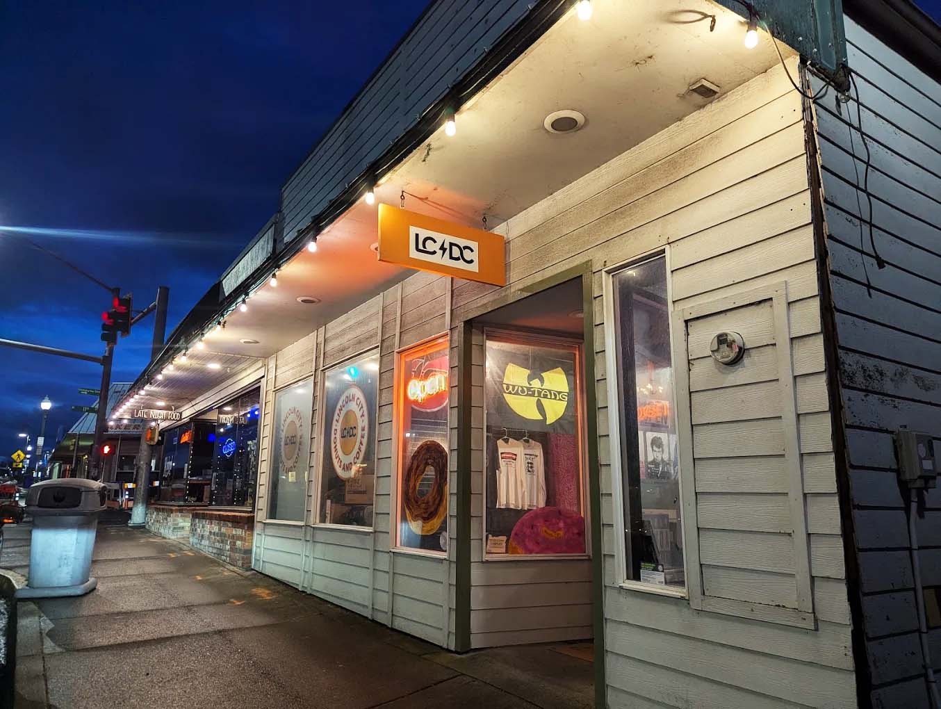 Photo by Jr. Velasquez via Google Local. The outside of Lincoln City Donuts And Coffee. There are rock and other music band logos in the windows, and the sign for the donut shop says LC DC, a play on the band AC/DC.