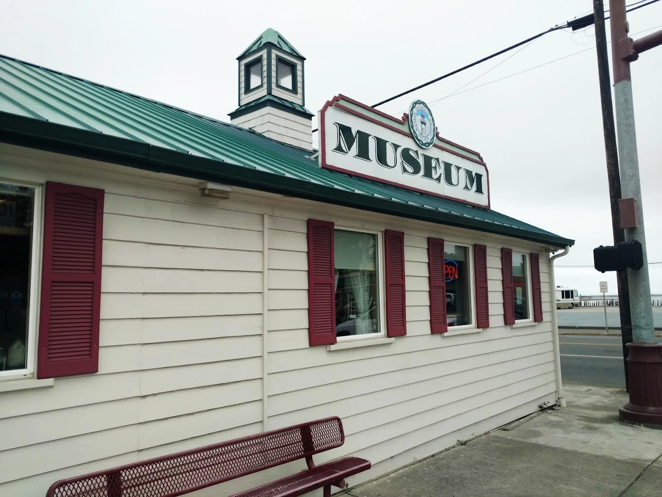 Photo by Kristopher Mullins via Google Local. The outside of the Bandon Historical Society Museum. It's a white building, with red shutters and a green roof.