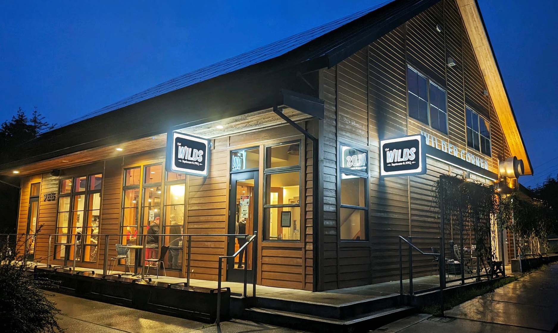 Photo via The Wilds Tap House & BBQ Facebook page. The outside of The Wild's Tap House & BBQ in Newport Oregon. It's a tall wood cabin style building, though modern. It had two black and white light up signs that say, 'The Wilds Taphouse & BBQ'. In the photo, it's night time, but a warm cozy glow emits from the windows and doors from inside the building.