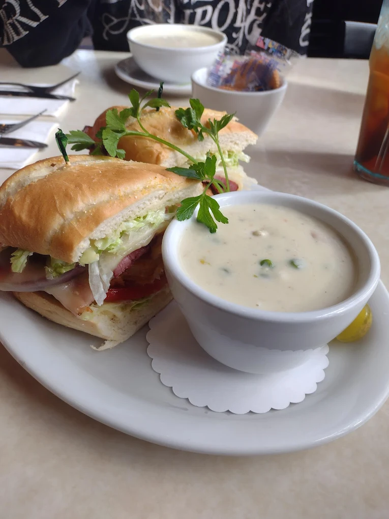 A thick sandwich on a roll with a bowl of white soup on a plate. It looks fancy.