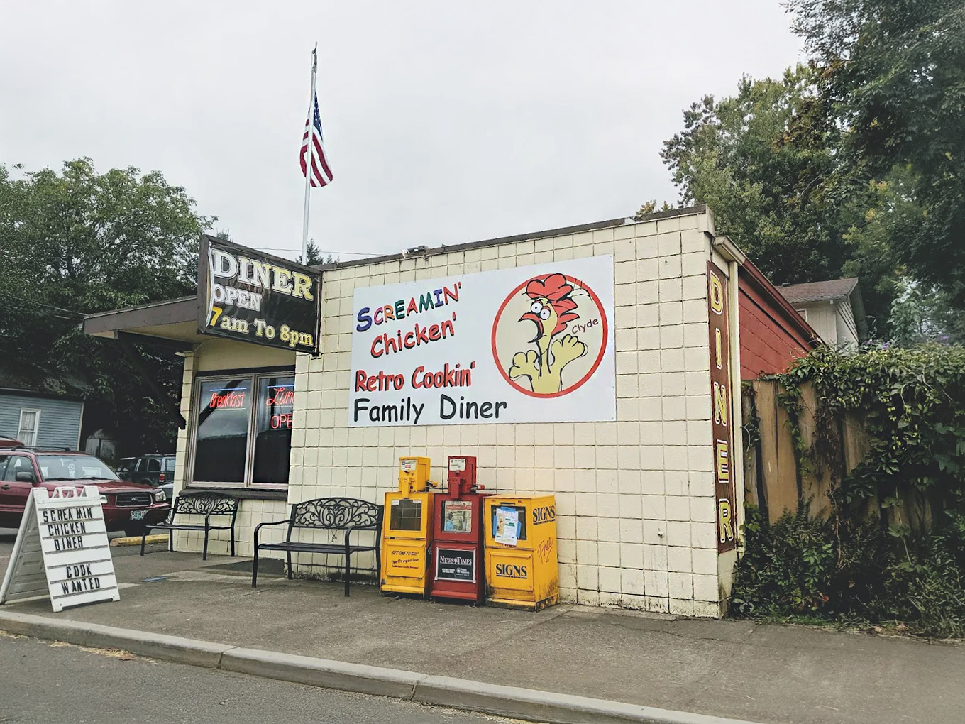 Photo by Akitomo Matsubayashi via Google Local. The outside of The Screamin' Chicken diner. It's a white building with a large sign of a screaming chicken. There are two black benches out in front on the sidewalk, and an American flag on a pole on the roof.