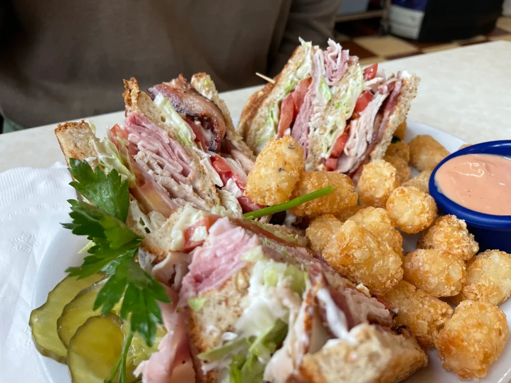 A crazy delicious looking sandwich. This thing is packed with meat and veggies and has a lot of layers to it. There are also tater tots and pickle slices on the plate. It looks delicious and really filling.