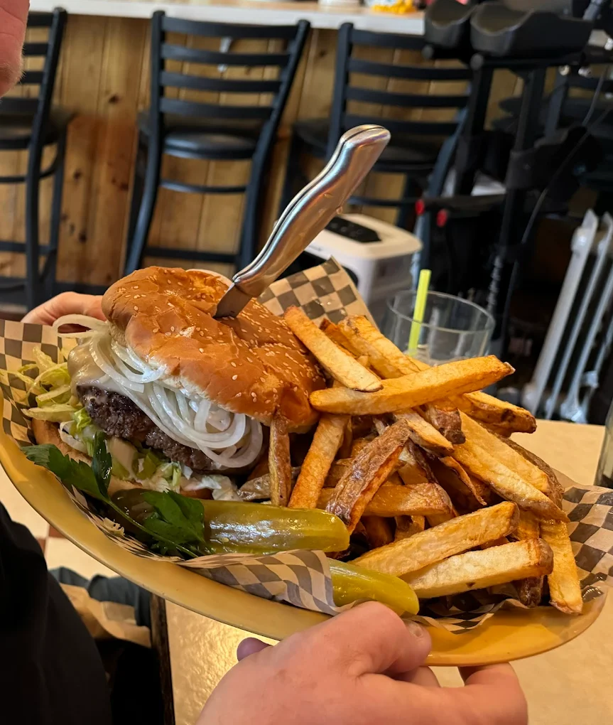 A crazy looking burger with a knife sticking out the top, large French fries and pickles on a plate with a black and white checkered food paper wrapper.