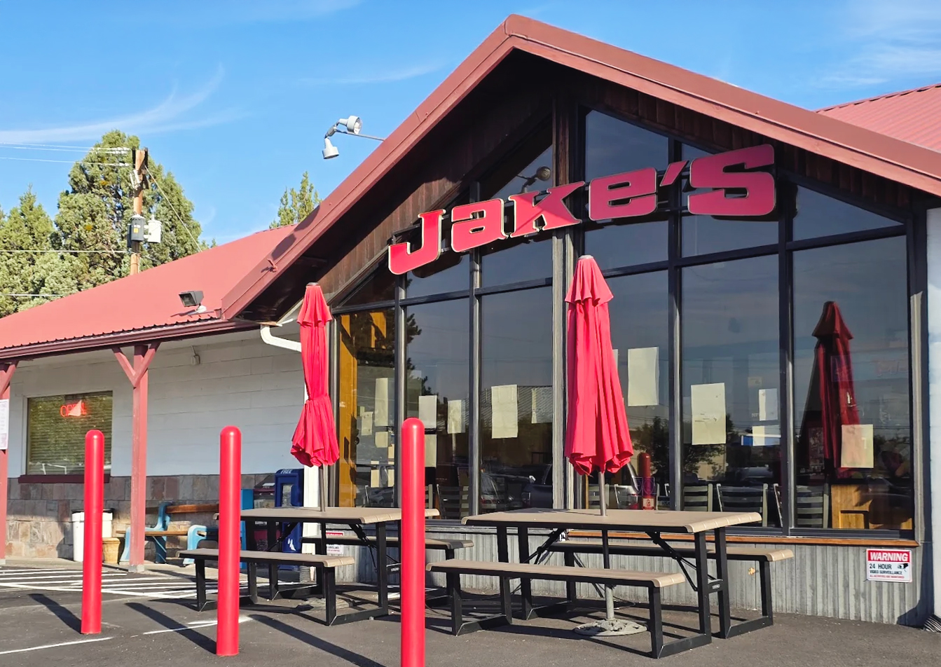 Photo by Mondo Gomez via Google Local. The outside of Jake's Diner in Bend, Oregon. It's a white building with red words, a reddish roof, large front windows, and other red accents around the exterior.