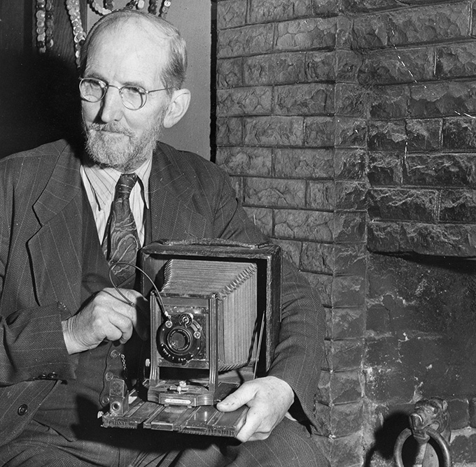 Historic black-and-white photo of June D. Drake seated beside his early 1902 view camera in 1954, a key figure in preserving Silver Falls State Park.